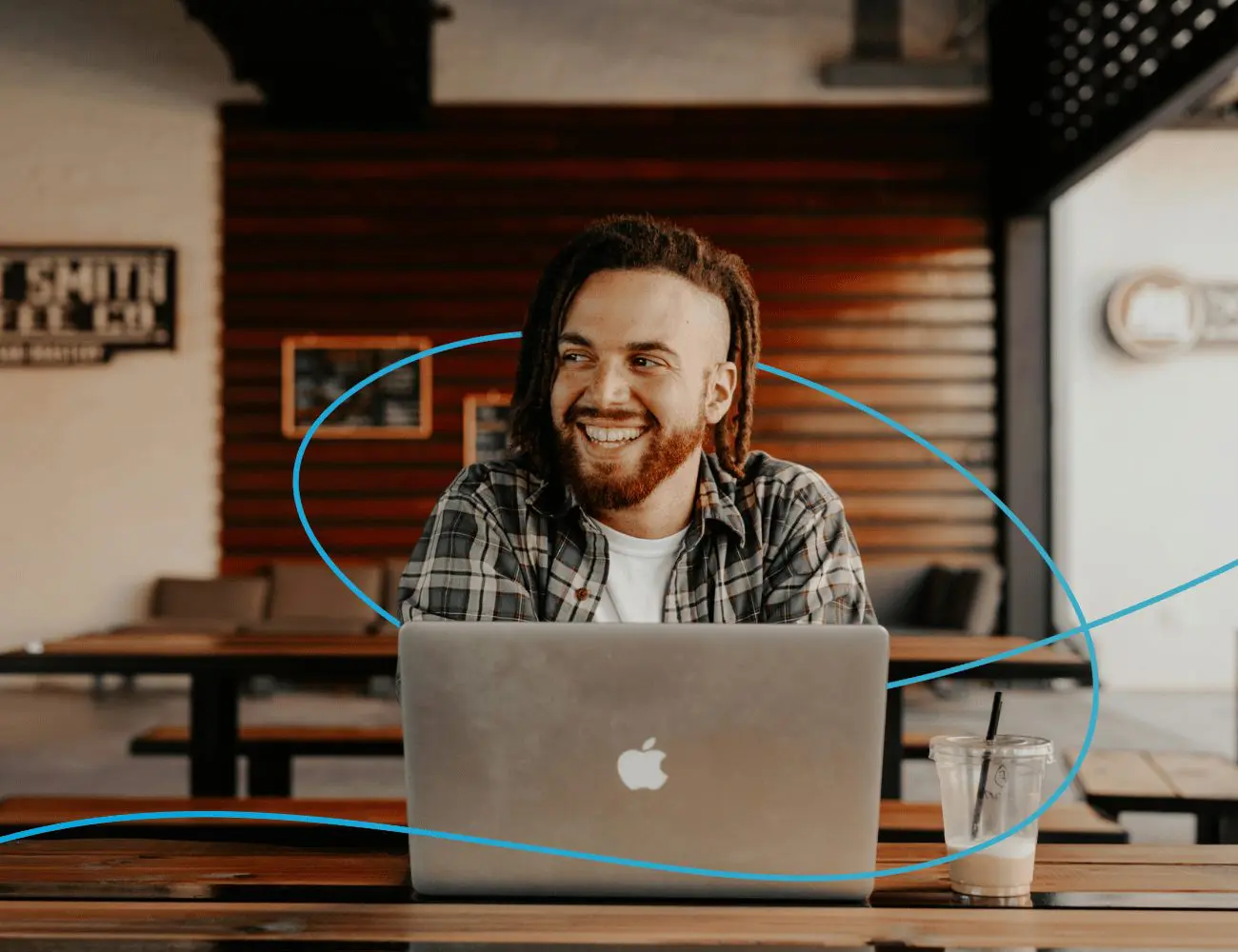 A young man is sitting at a table in a café, laughing and working on a laptop.
