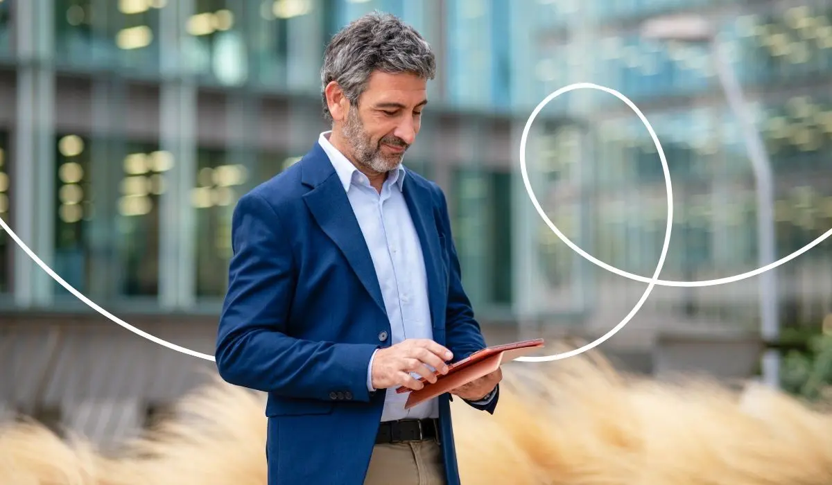 Älterer Mann in Business-Outfit steht vor einem großen, verglasten Bürogebäude und tippt auf einem Tablet. Symbolbild für die Arbeit mit einem eigenen CRM bzw. Loyaltyprogramm.