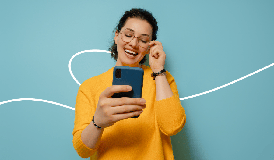 Kobieta w pomarańczowym swetrze stoi na neutralnym niebieskim tle i patrzy na smartfon. Śmieje się przy tym i dotyka swoich okularów.