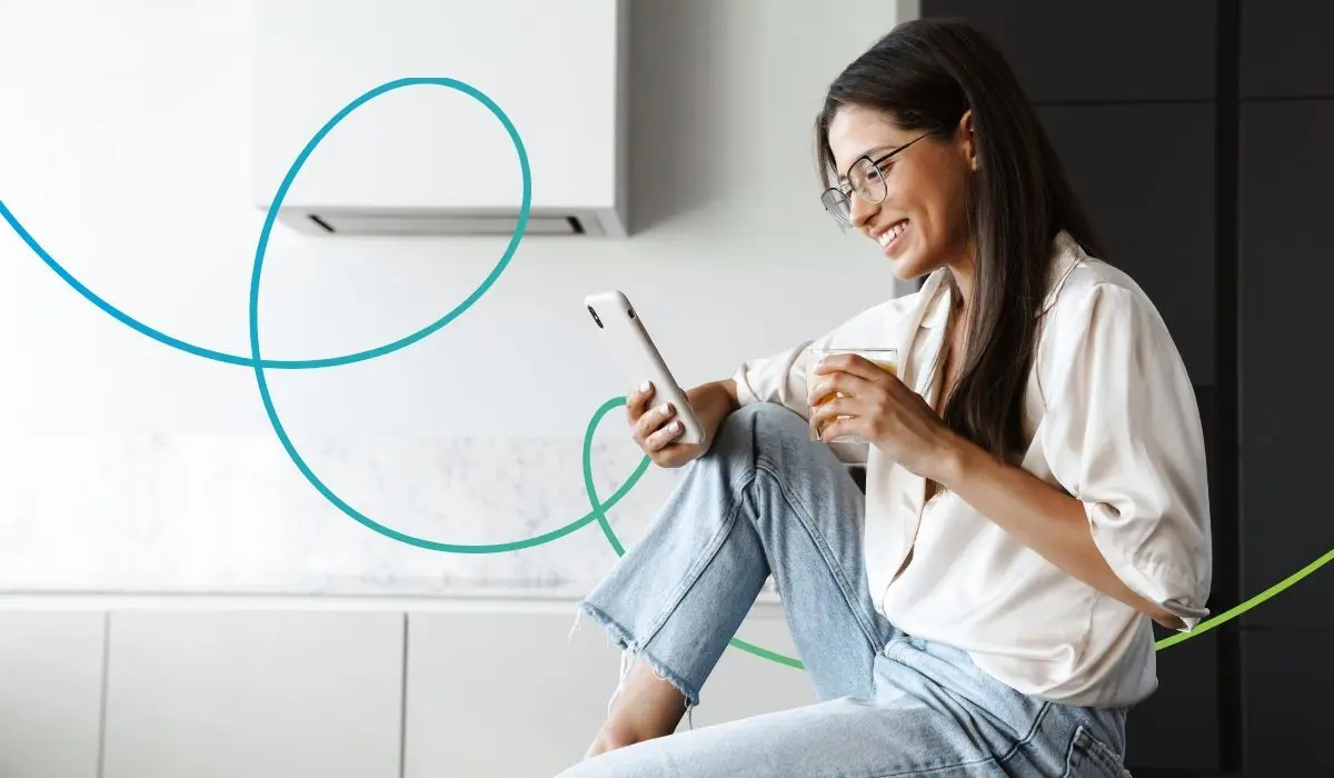 Junge frau sitzt in einem Wohnzimmer mit einem Smartphone in der Hand. Sie schaut lächelnd auf das Handy.