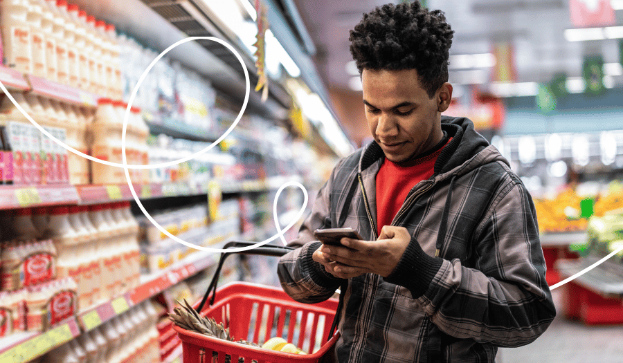 A man is standing in front of a refrigerated display case in a supermarket, looking at his smartphone.