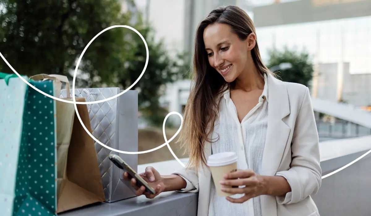 A woman holding a coffee-to-go cup looks at her cell phone in her other hand and logs into a loyalty app with her information. Shopping bags are in the background.