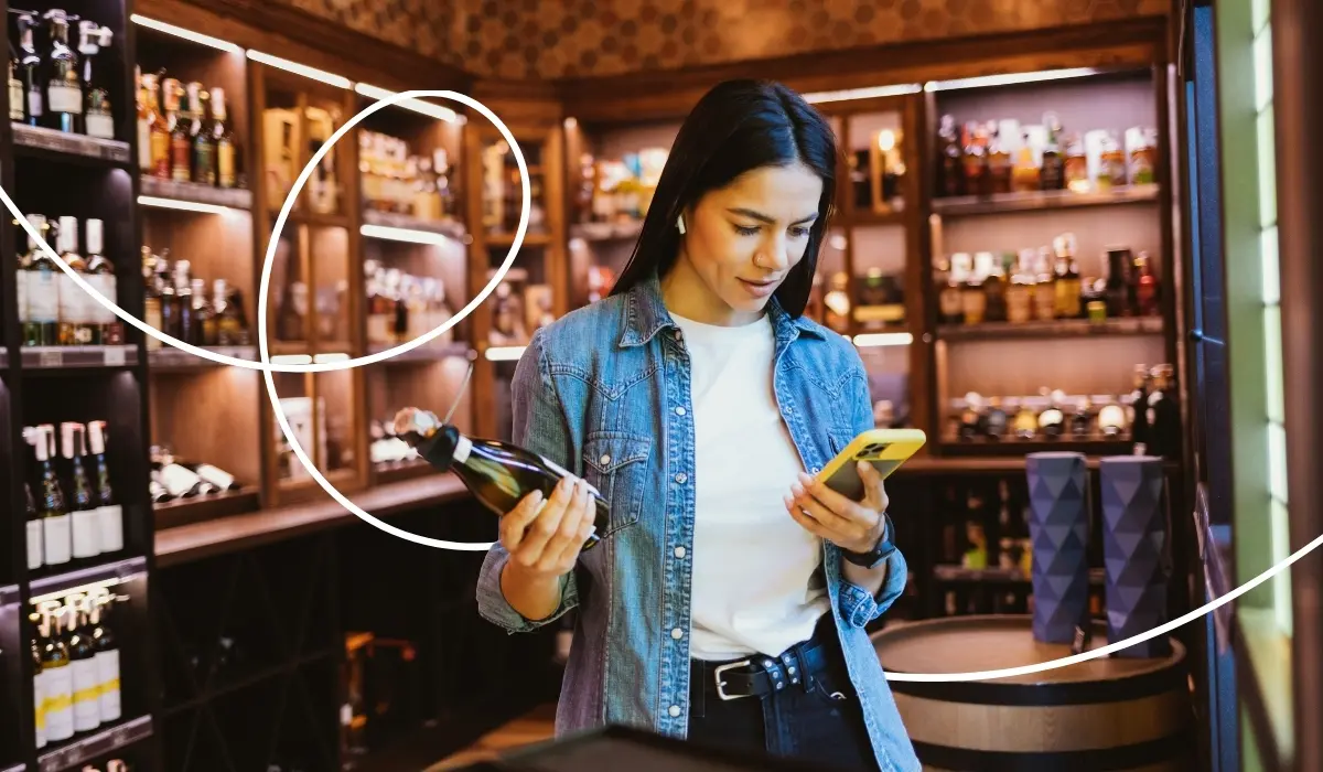 A young woman is standing in a wine shop, searching for discount offers on her cell phone.