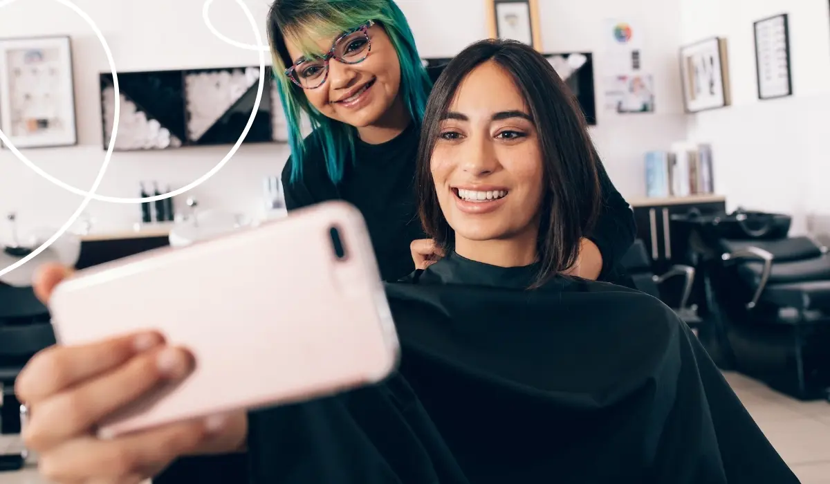 Eine Friseurin und eine Kundin sind in einem Friseursalon und schauen gemeinsam auf ein Kundenbindungsprogramm auf dem Smartphone.