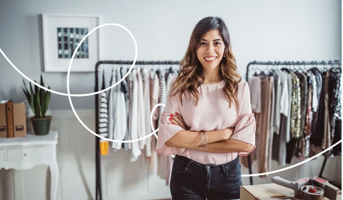 Modehändlerin steht in ihrer Boutique und schaut freundlich mit verschränkten Armen in die Kamera.