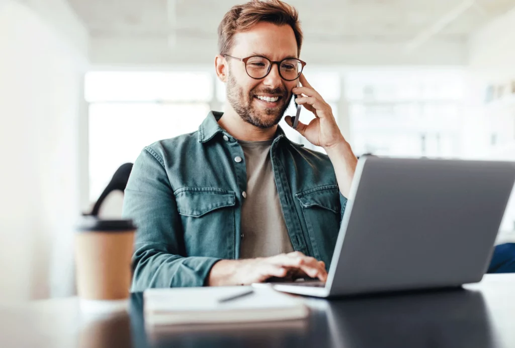 Junge Mann sitzt an einem Tisch und arbeitet mit einer Hand am Laptop, während er mit der anderen telefoniert. Er lächelt und neben ihm steht ein Becher Kaffee.