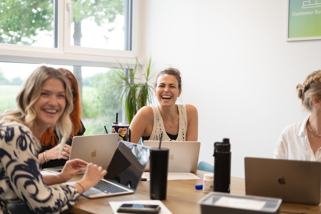 Twee medewerksters van hello again zitten samen met andere collega's in een vergaderruimte achter hun laptop en lachen naar de camera.