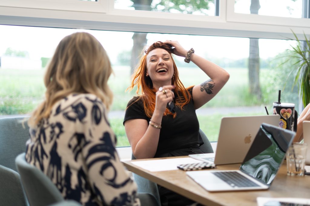 Een medewerkster van hello again zit samen met andere collega's in een vergaderruimte bij hello again en lacht.